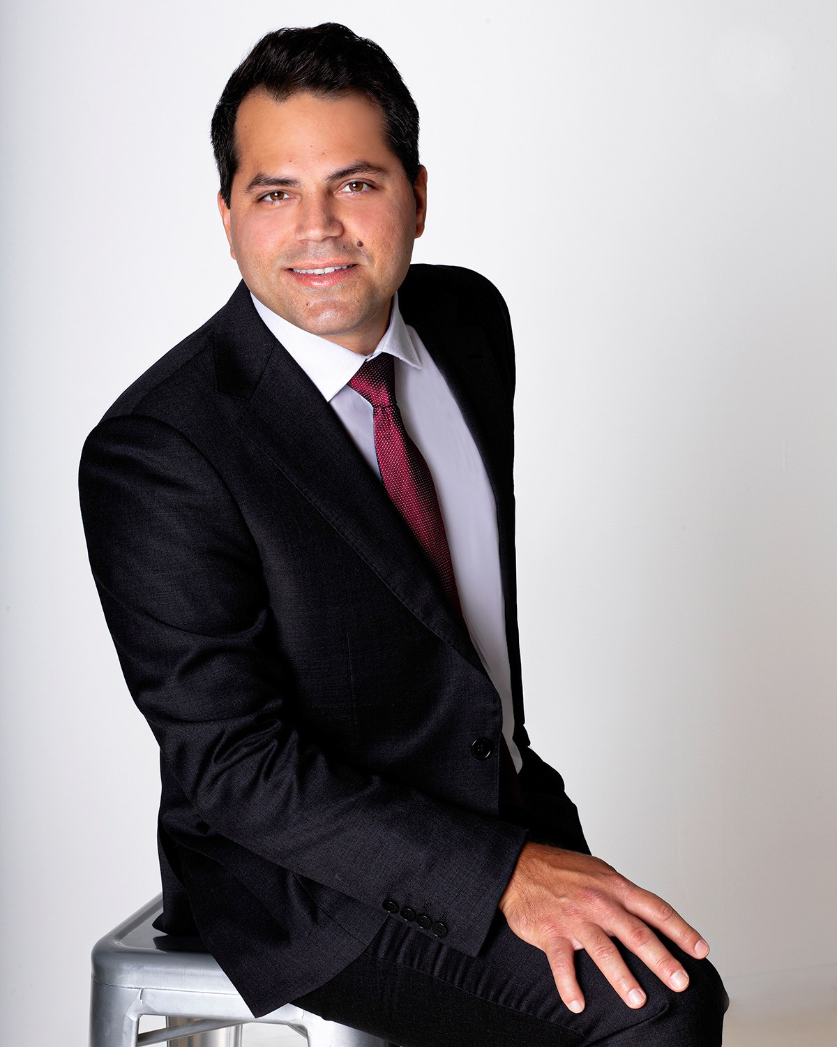 Man in a dark suit and burgundy tie, seated and smiling, white backdrop, photographed by Annie Watson in her Naples studio